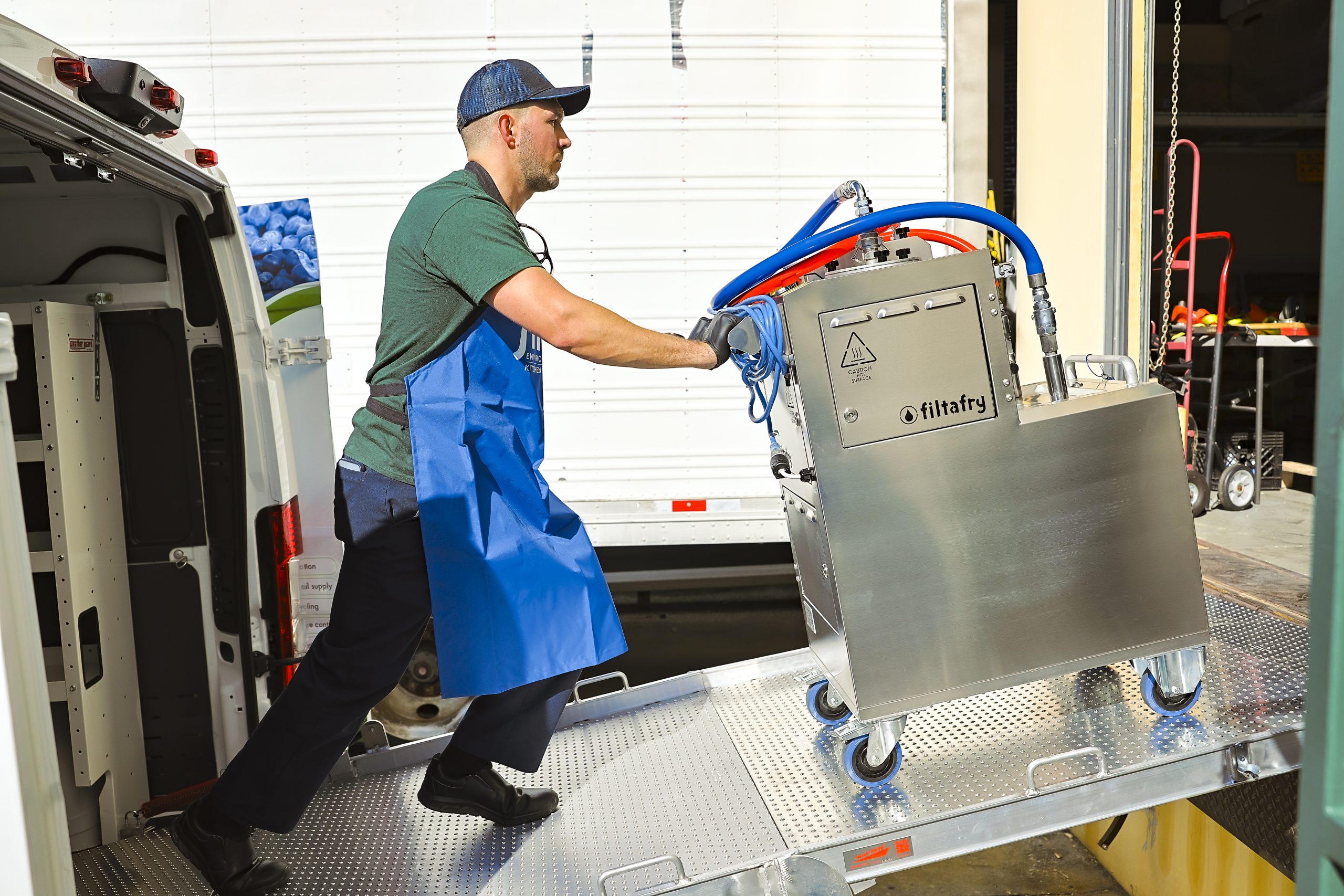 A person in a hat and blue uniform pushing an cooking oil filtration unit up a ramp.